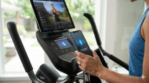 A detailed, photorealistic 4K image based on image_0.png, focusing on the elliptical console and the user's hand holding a smartphone, which displays a pulsing Bluetooth pair icon to illustrate syncing with a fitness app. In the background, the tablet still shows the workout class.